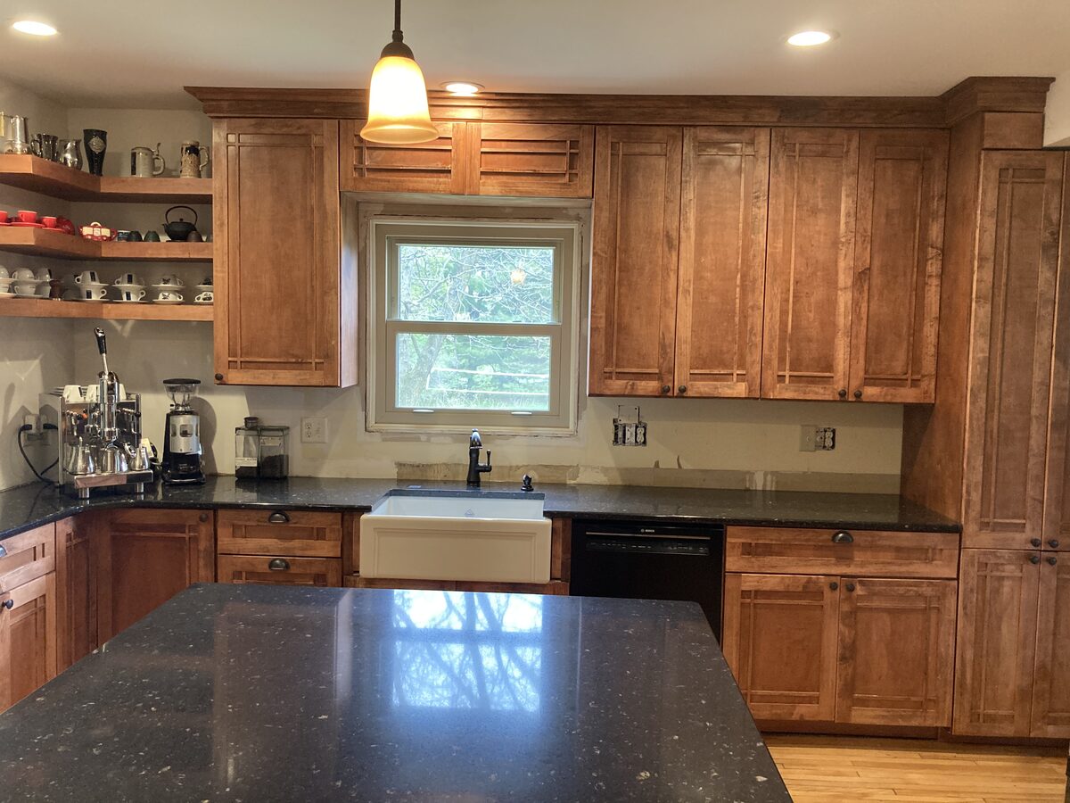 Wide view of craftsman kitchen remodel with brown cabinets, farmhouse sink, open shelving, and black countertops