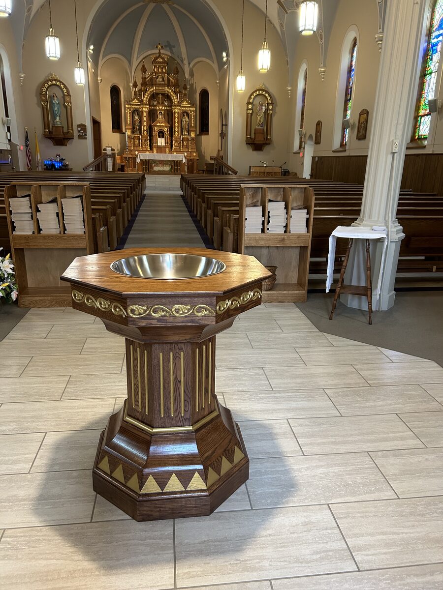 Custom baptismal font installed in a Catholic church with ornate golden altar and stained glass windows visible down the center aisle